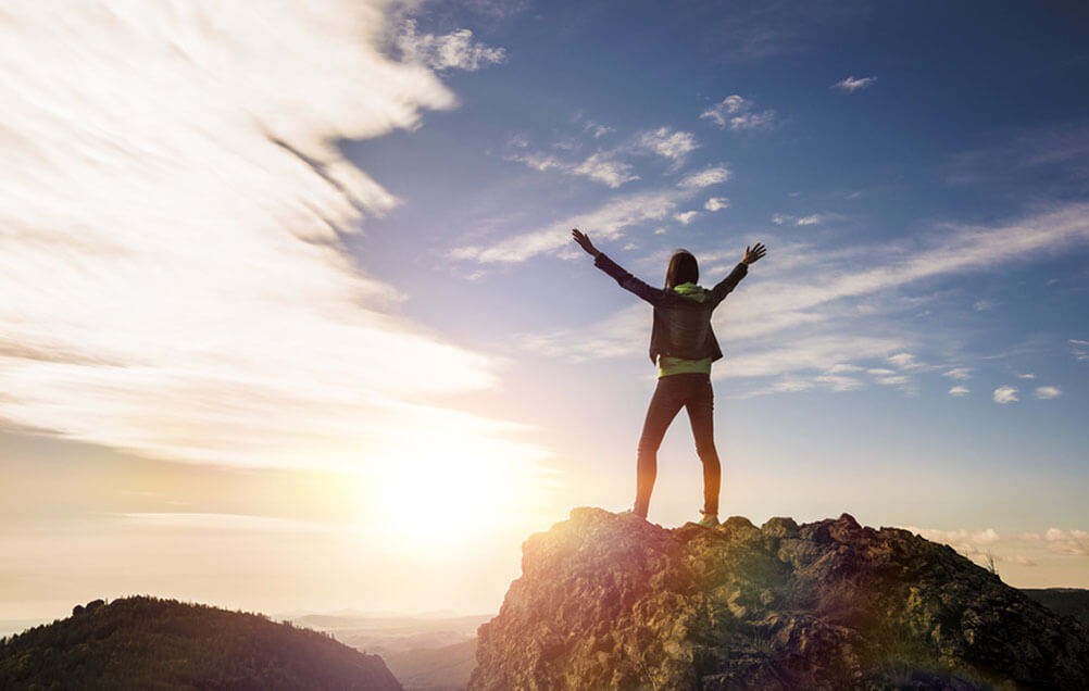 Against the Odds Woman stands on top of a mountain with arms outstretched as the sun sets