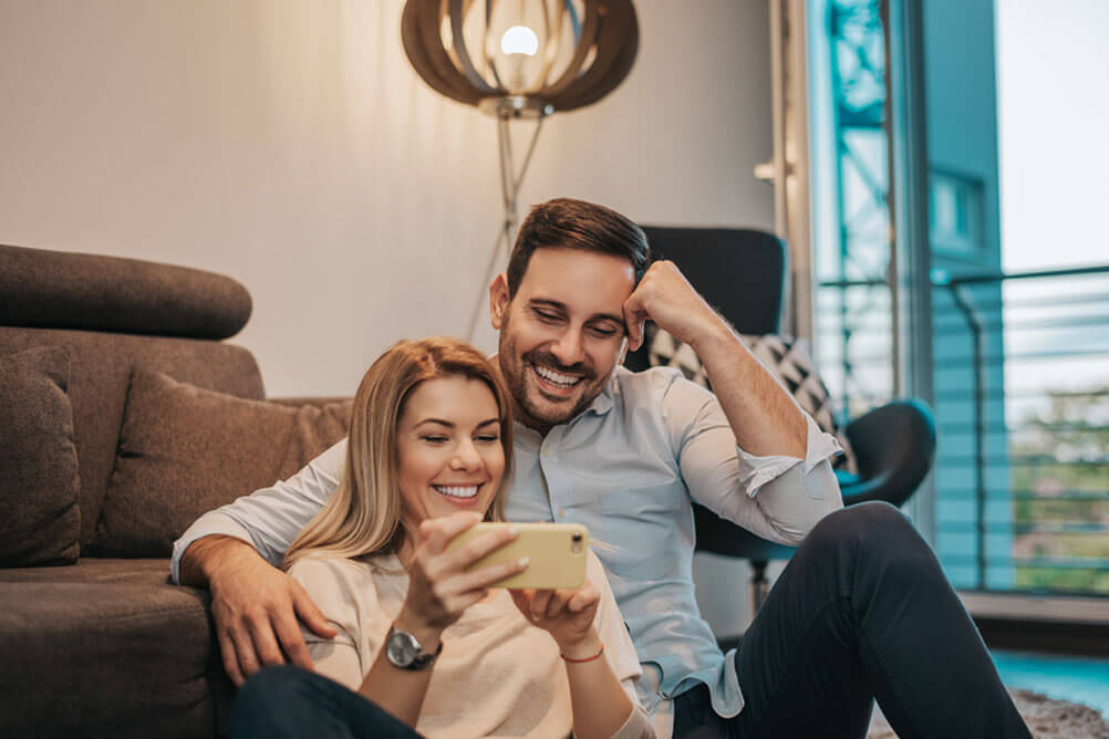 Man and woman seated on floor in front of sofa looking at smartphone and smiling