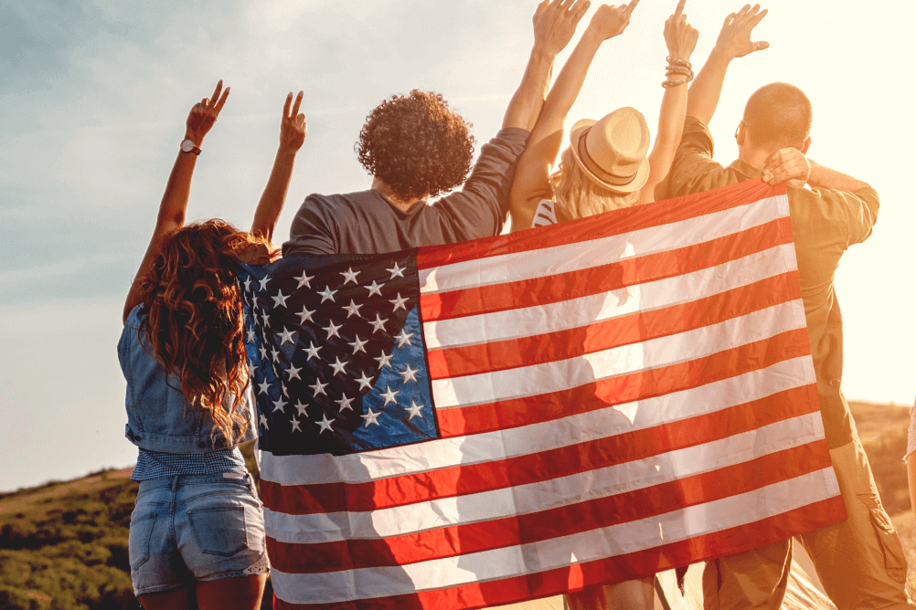 Group of four friends outdoors waving while carrying the American flag