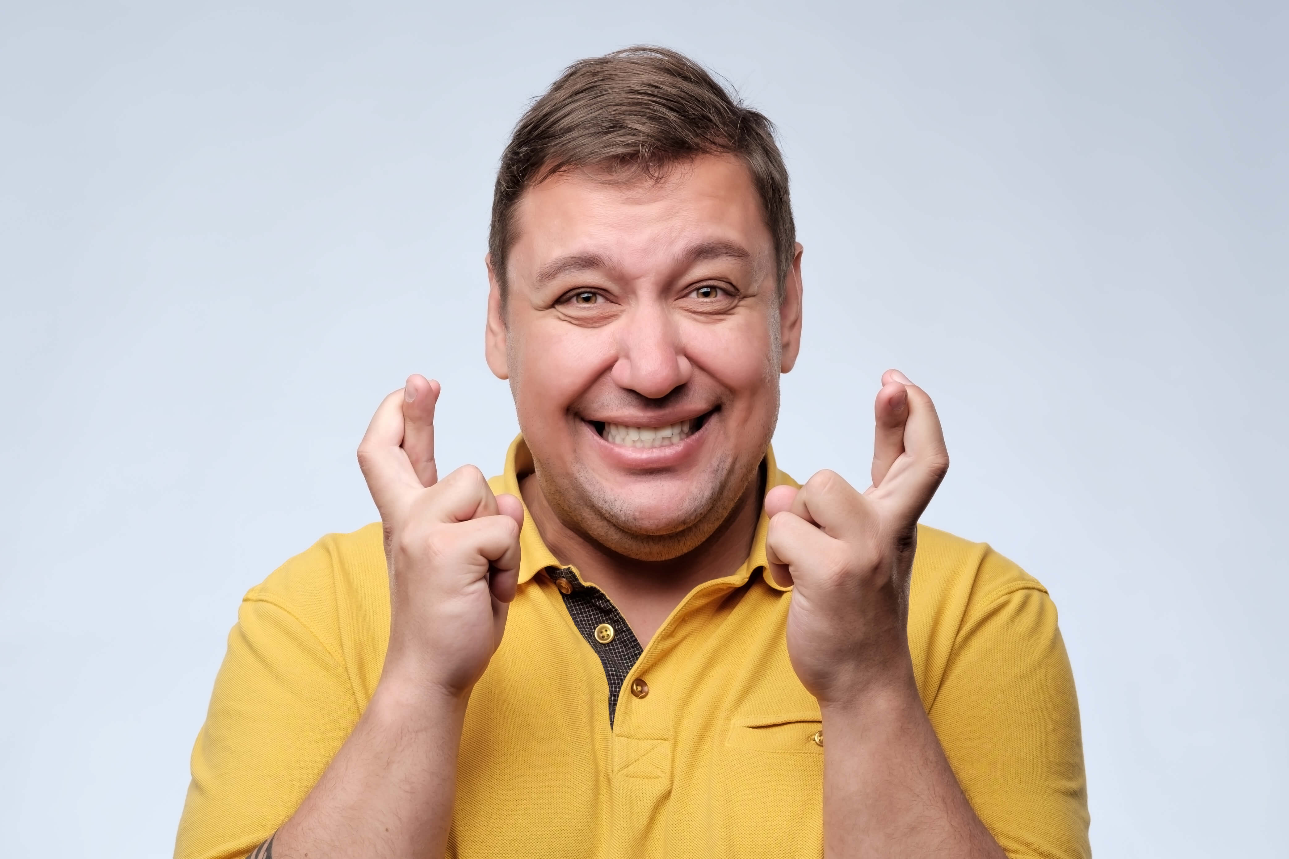 Smiling man in a yellow shirt crosses his fingers in the hope of getting good luck