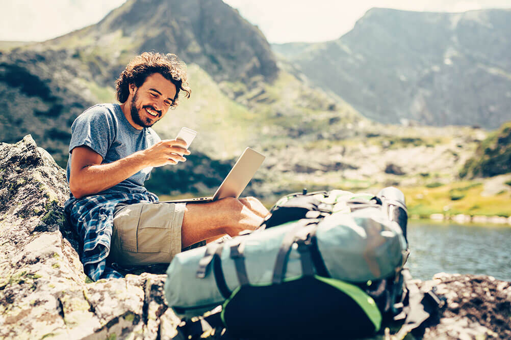How to Become a Digital Nomad A man smiles as he works as a digital nomad from a beautiful location next to mountains and water